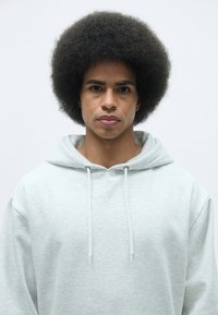 Felpa grigia chiara con cappuccio e coulisse, morbida al tatto, vestibilità ampia, e un grande afro in primo piano su uno sfondo neutro.