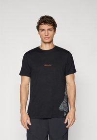 Man met krullend haar in een zwart T-shirt met korte mouwen, voorzien van de tekst "icebreaker" en een contourpatroon aan de zijkant, staand tegen een effen achtergrond.