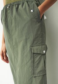 Olivgrüner Cargo-Rock mit elastischem Bund, Metall-Schnapp-Taschen und einer Hand in der rechten vorderen Tasche.