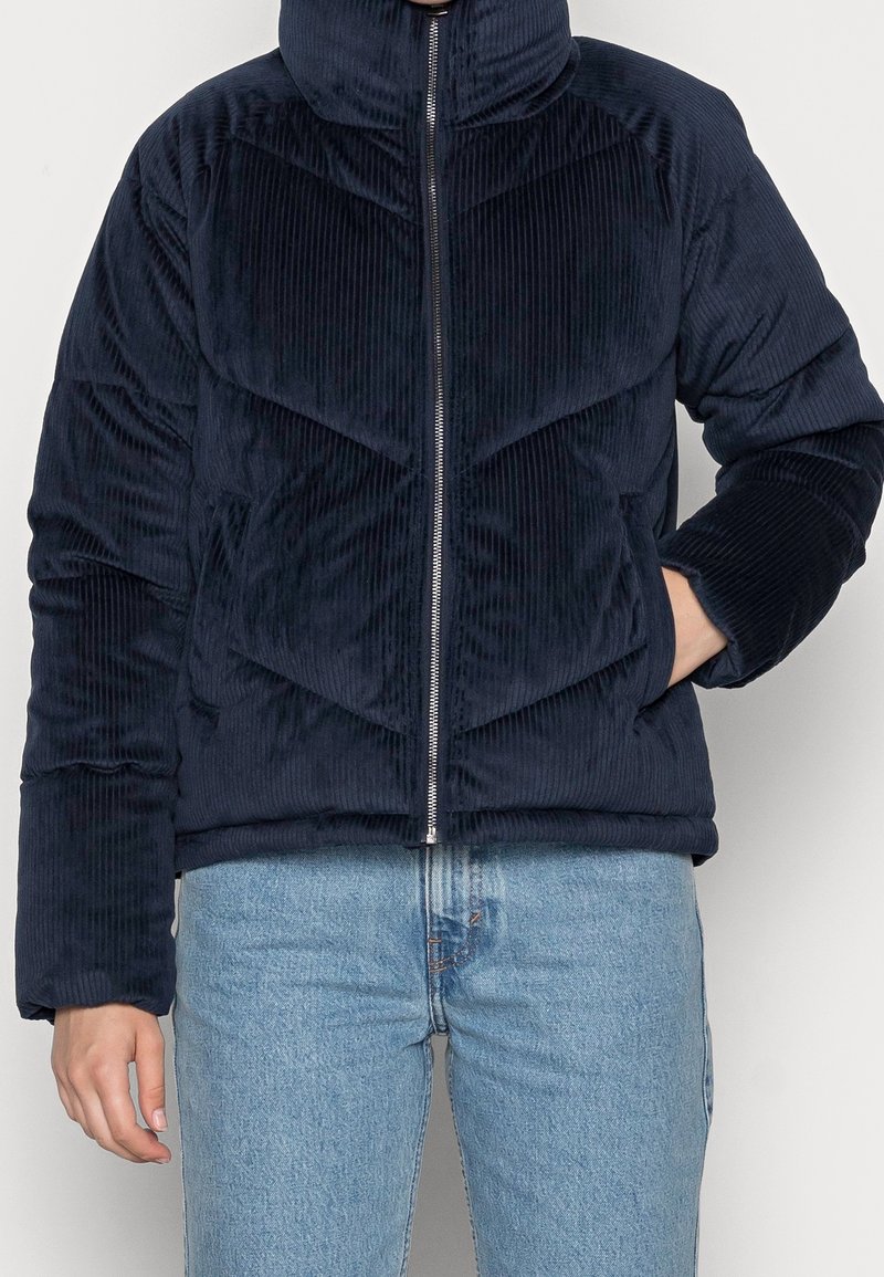 Granatowa kurtka puchowa z kordury z wysokim kołnierzem, pionową fakturą, poziomym przeszyciem w kształcie litery V i zamkiem błyskawicznym z przodu.