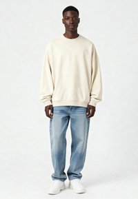 Bežinis per didelis džemperis su raukšlėtu apykakle, derinamas su šviesiai mėlynomis tiesiomis kelnėmis ir baltomis sportinėmis bateliais, neutralios spalvos fone.