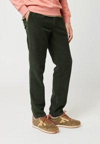 Man in donkergroene corduroybroek, tan en bruine sneakers met oranje veters, en een zalmroze sweatshirt, staande tegen een witte achtergrond.