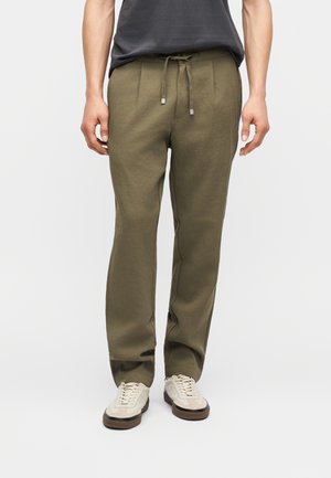 Άνδρας που φοράει χακί παντελόνι με κορδόνι και μπέζ αθλητικά παπούτσια, στέκεται μπροστά σε απλό ανοιχτόχρωμο φόντο.