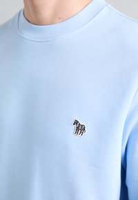 Heledsinine pehme teksapusa, millel on rinnaosa väike, värvikas tikand hobuse kujutisega. Ümmargune kaelus, juhuslik disain.