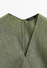 Vêtement en tissu vert texturé avec un col en V et une étiquette noire indiquant Massimo Dutti cousue à l’intérieur de l’encolure.