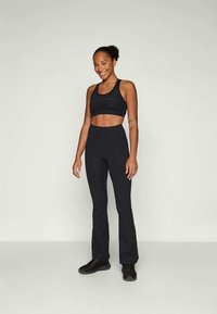 Soutien-gorge de sport noir et pantalon taille haute, design ajusté avec une texture lisse, porté avec des baskets noires. Le modèle se tient devant un fond uni.