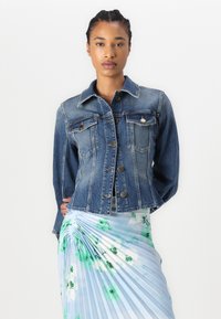 Denim jacket featuring a fitted design, light blue color, button front, and two chest pockets. The model wears a pleated floral skirt.