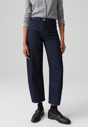 Personne portant un jean droit taille haute bleu marine, un pull gris boutonné, des chaussettes transparentes noires et des mocassins en cuir noirs sur un fond uni.