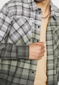 Rutig skjorta i gröna och grå nyanser, med knäppning fram, bröstficka och långa ärmar med manschetter. Slät textur, neutral baslager.