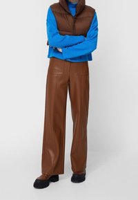 Pantaloni larghi in finta pelle marrone con tasche frontali, abbinati a un maglione blu e a un gilet imbottito marrone. Scarpe chunky nere completano il look.