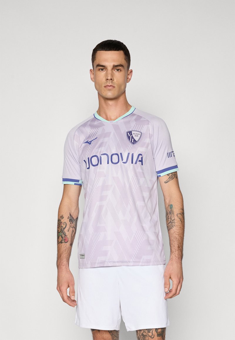 Homme portant un maillot de football VfL Bochum violet avec des logos de sponsors et un short blanc, se tenant contre un fond uni.
