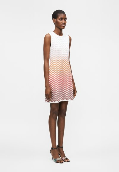 Γυναίκα φοράει άσπρο αμάνικο φόρεμα με πορτοκαλί και ροζ σχέδιο ζικ-ζακ, συνδυασμένο με μαύρα σανδάλια με ψηλό τακούνι, στέκεται σε απλό φόντο.