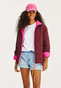 Burgundská prošívaná bunda s růžovými akcenty, spárovaná s bílým tričkem a džínovými šortkami. Bunda má zip a barevné šňůrky.
