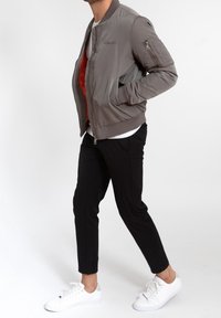 Man wearing grey bomber jacket, white sneakers, black cropped pants, and white shirt, standing against a plain white background.