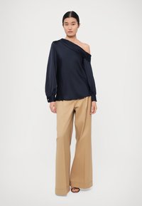 Navy off-shoulder blouse met een vloeiende silhouet en lange, geplooide mouwen, gecombineerd met beige wijde broek en sandalen.