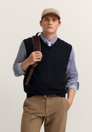 Black sleeveless sweater over a blue and white striped shirt, light brown belt, khaki pants, and a beige cap, holding a brown shoulder bag.