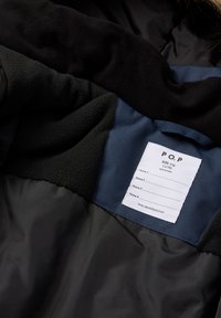 Binnenkant van een zwarte en marineblauwe jas met een wit kledinglabel waarop maat, leeftijd en ruimte voor vier namen staan.