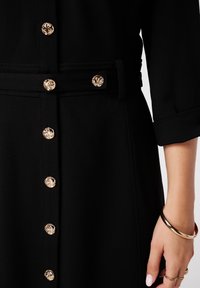Zwarte jurk met gouden decoratieve knopen aan de voorkant, een stoffen riemdetail en driekwartmouwen. Soepele textuur.
