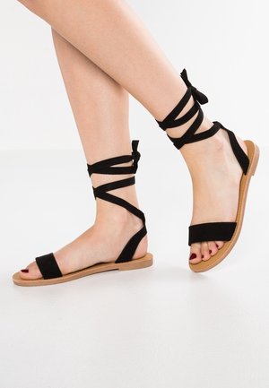 Zwarte suède sandalen met dunne enkelbandjes, platte zolen, een brede te band en een contrasterende beige zool voor extra detail.