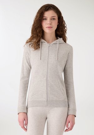 Giovane donna con capelli ricci che indossa una felpa con cappuccio grigio chiaro con cerniera e pantaloni abbinati, in piedi davanti a uno sfondo bianco semplice.