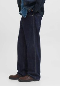 Vaqueros de mezclilla azul oscuro con un corte holgado, con costuras amarillas, bolsillos laterales y un diseño de pierna recta. Combinados con zapatos marrones.