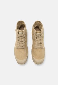 Zapatillas altas beige de lona con acabado texturizado, que presentan un diseño de cordones y una puntera de goma, mostrando una etiqueta en la lengüeta.