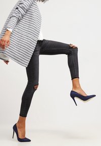 Persona che indossa jeans skinny neri strappati, tacchi alti blu, e un blazer rigato grigio e bianco a metà passo su uno sfondo semplice.
