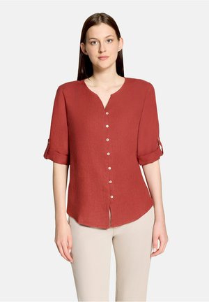 Vrouw draagt een roestrode blouse met knopen en opgerolde mouwen en beige broek, staand tegen een effen lichte achtergrond.