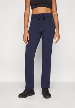 Kvinde iført mørkeblå joggingbukser med bindebånd i taljen, sort crop top og sorte sneakers står mod en ensfarvet lys væg.