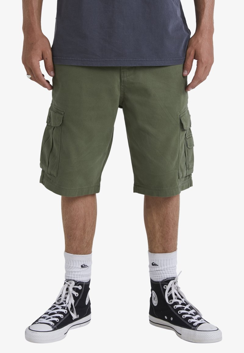 Green cargo shorts with multiple side pockets, made from durable fabric. Worn with black high-top sneakers and white crew socks.