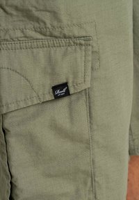 Pantalones cortos de carga verdes hechos de tejido ligero, con un bolsillo lateral y una pequeña etiqueta negra con texto blanco en la cintura.