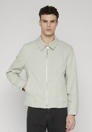 Ung mand iført lysegrå lynlåstrøje med subtilt teksturmønster og sorte bukser, stående med den ene hånd i lommen.