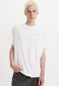 Camiseta de algodón blanca con mangas cortas, cuello redondo y un sutil logo gris de Levi's en el lado izquierdo del pecho. Diseño simple y corte relajado.