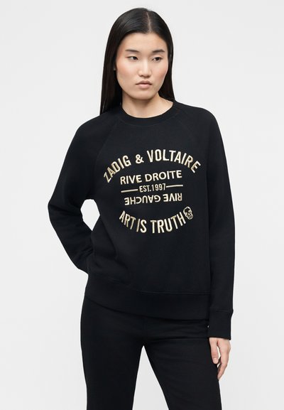 Γυναίκα με ίσια μαύρα μαλλιά, φοράει μαύρο φούτερ με μπεζ κείμενο «Zadig & Voltaire Rive Droite Est. 1997 Art is Truth», στέκεται μπροστά σε απλό φόντο.