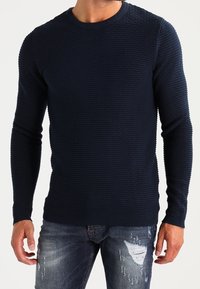 Pull en tricot côtelé bleu marine avec un col rond, des manches longues et un motif texturé. Associé à un jean en denim déchiré.