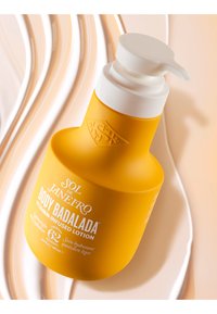 Gelbe Flasche mit Körperlotion und weißem Pumpdeckel, mit erhabenem Logo und bedrucktem Etikett, das den Produktnamen und die Beschreibung enthält.