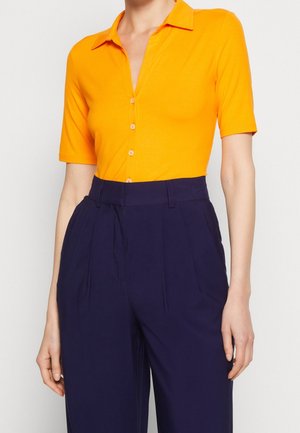 Persoon draagt een fel oranje korte mouw overhemd met knopen, ingestopt in hooggetailleerde, donkerblauwe geplooide broek.