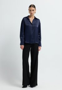 Donna in piedi contro uno sfondo bianco, che indossa una camicia satinata blu navy con bottoni, pantaloni larghi neri, tacchi neri e orecchini a cerchio dorati.
