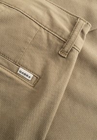 Közelkép bézs színű, texturált anyagból készült nadrágról, amelyen hátul egy „GABBA” feliratú zseb és egy övhurkó látható.