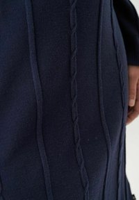 Tumesinine kudumisel riie, millel on kõrgendatud lõngakudumise mustrid ja sile tekstuur, sobituv siluett ja mugav käte asetamise koht.