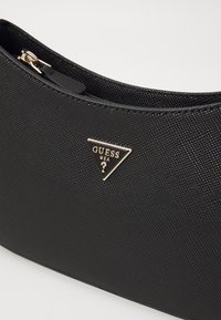 Sac à main noir texturé avec une fermeture éclair dorée et un logo triangulaire doré Guess USA à l'avant.