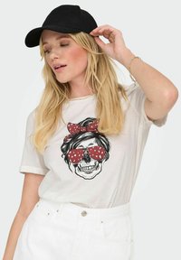 Camiseta blanca con un gráfico de calavera que lleva gafas de sol de lunares rojos y un lazo a juego, combinada con pantalones cortos blancos de cintura alta y una gorra negra.