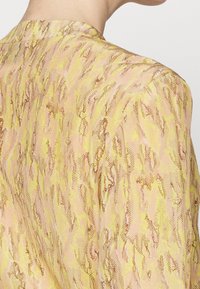 Tissu à motif imprimé peau de serpent jaune et marron sur l'épaule et le haut du dos d'un vêtement, porté par une personne aux cheveux courts.