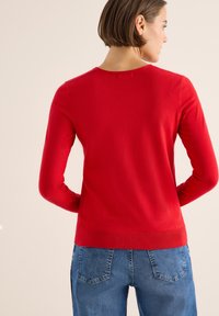 Roter, langärmliger Pullover mit rundem Halsausschnitt, tailliertem Schnitt und geripptem Saum. Kombiniert mit hochgeschnittenen blauen Jeans.