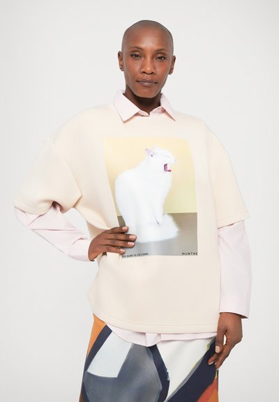 Svetlo roza oversize pulover z grafiko bele mačke, z gladko teksturo in okroglo ovratnikom, položen preko roza srajce.