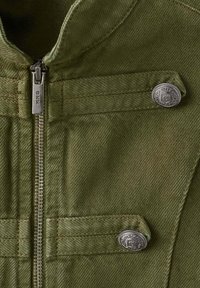Tissu vert olive avec fermeture éclair verticale et deux sangles horizontales maintenues par des boutons métalliques argentés décoratifs avec un dessin de blason gravé.