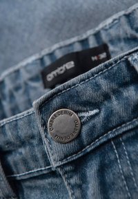 Nærbillede af blå denimjeans med fokus på en metallisk knap mærket "OMBRE DENIM" og et sort indvendigt mærke.