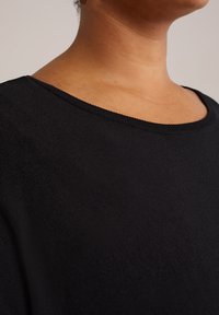 Gros plan sur le cou et le haut de la poitrine portant un haut en tricot noir à col rond, sur un fond neutre.