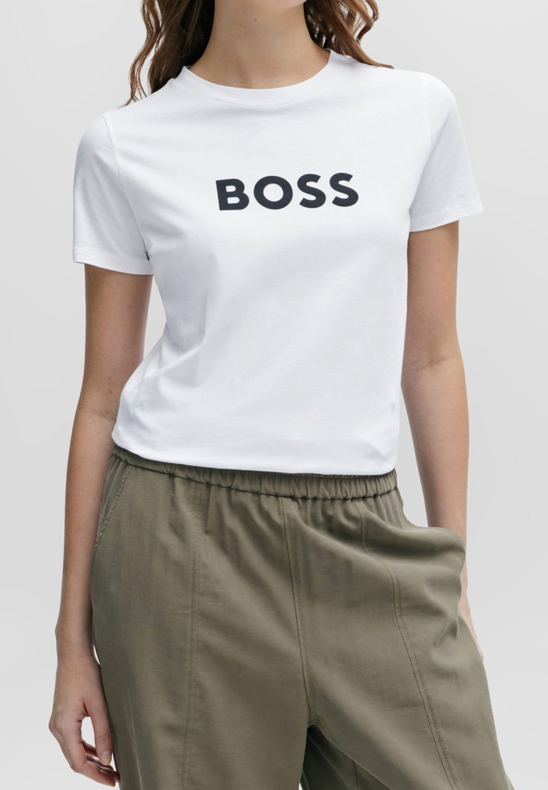 Vit bomullströja med "BOSS" tryckt i fet svarta bokstäver. Korta ärmar, rund halsringning, enkel design, kombinerad med olivgröna byxor.