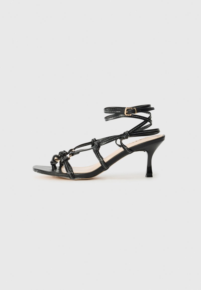 Black patent leather high-heeled sandal with strappy design, adjustable ankle strap, and knotted detailing. Smooth texture, pointed toe.